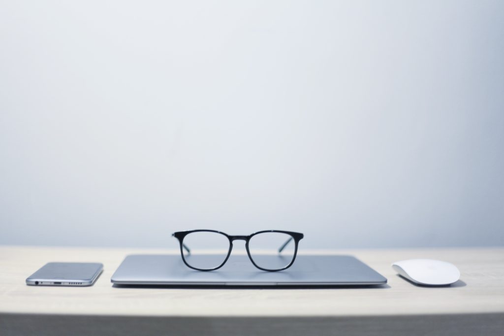A pair of black glasses sit on top of a small laptop next to a phone and a mouse