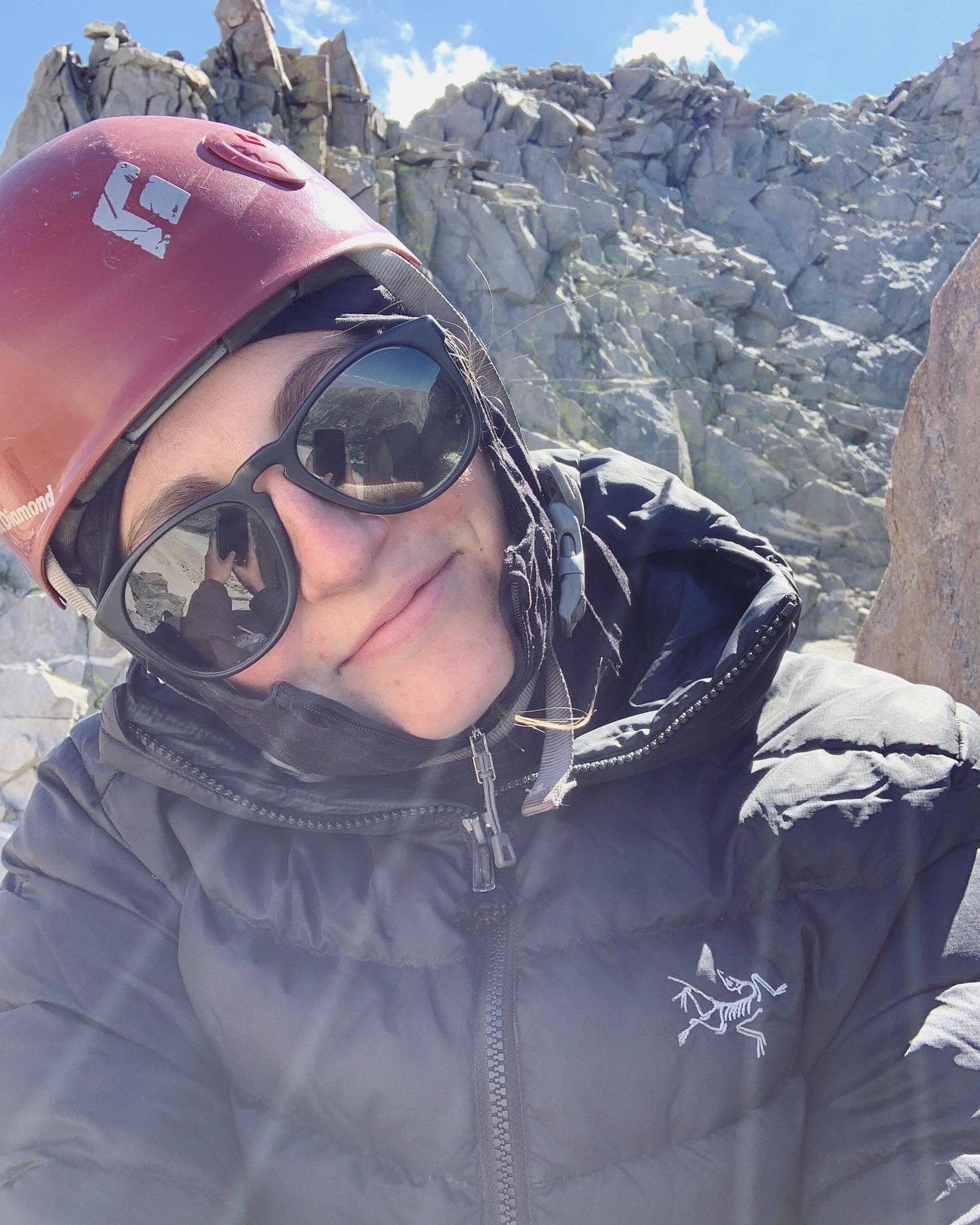 A selfie of Jessica wearing a rock climbing helmet, sunglasses and a black puffy jacket taken while climbing an alpine rock route