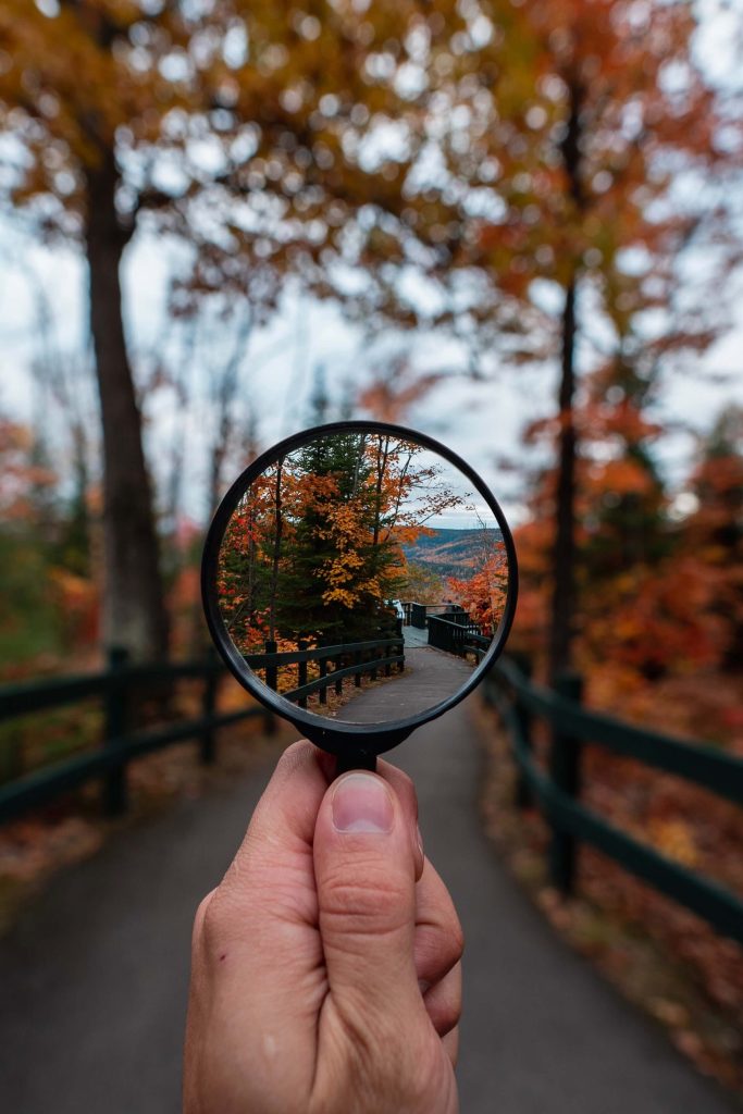 A hand holds a magnifying glass in the foreground of a paved trail with split rail fence on both sides and bright orange fall foliage surrounding the trail